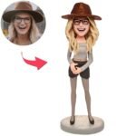 Cartoon woman in a brown hat, glasses, gray top and black shorts, hands clasped, on a round base; inset shows the real person with the same hat.