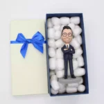 Open gift box with blue ribbon revealing a businessperson bobblehead nestled in packing peanuts
