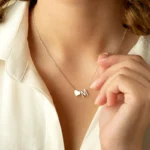 Close-up of a woman wearing a delicate gold necklace with a small heart charm and the letter “M” pendant. She is holding the necklace lightly with her fingers, showcasing a personalized initial jewelry design against a soft white blouse.