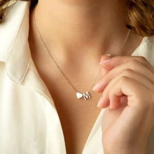 Close-up of a woman wearing a delicate gold necklace with a small heart charm and the letter “M” pendant. She is holding the necklace lightly with her fingers, showcasing a personalized initial jewelry design against a soft white blouse.