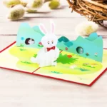 3D Easter pop-up greeting card featuring a cute white bunny standing in a colorful spring meadow scene with green hills, flowers, and Easter decorations. A handmade paper card design for Easter greetings, seasonal gifting, and festive celebrations.