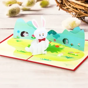3D Easter pop-up greeting card featuring a cute white bunny standing in a colorful spring meadow scene with green hills, flowers, and Easter decorations. A handmade paper card design for Easter greetings, seasonal gifting, and festive celebrations.