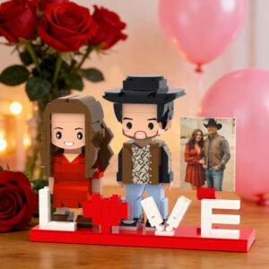 A personalized LEGO-style couple figurine display on a red base spelling “LOVE,” with a heart replacing the letter “O.” Two block-style figures—a woman in a red dress and a man wearing a hat—stand beside the word. A small framed photo of the real couple is placed next to them, with romantic decorations including red roses, pink balloons, and warm ambient lighting in the background.
