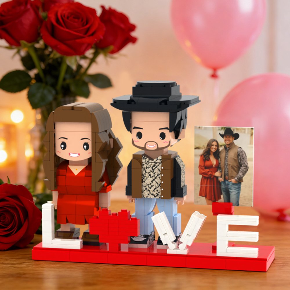 1fd5781e-67db-4b9f-bd11-d8878f5c801c-0jpeg - printshop4me.ae A personalized LEGO-style couple figurine display on a red base spelling “LOVE,” with a heart replacing the letter “O.” Two block-style figures—a woman in a red dress and a man wearing a hat—stand beside the word. A small framed photo of the real couple is placed next to them, with romantic decorations including red roses, pink balloons, and warm ambient lighting in the background.