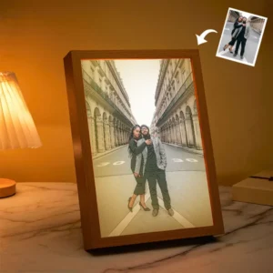 Framed personalized couple photo displayed on a tabletop under warm ambient lighting, featuring a stylized portrait of a couple standing together on a street between classical buildings. A small reference photo appears in the top-right corner indicating the original image used for the custom artwork transformation, suggesting a photo-to-portrait frame gift for couples or anniversaries.