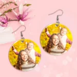 Two round personalized photo earrings with silver hooks displayed on a pink background with floral accents. Each earring features a bright outdoor photo of a smiling mother and child in warm autumn scenery, designed as custom printed photo jewelry gifts.