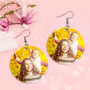 Two round personalized photo earrings with silver hooks displayed on a pink background with floral accents. Each earring features a bright outdoor photo of a smiling mother and child in warm autumn scenery, designed as custom printed photo jewelry gifts.