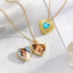 Two gold heart-shaped lockets displayed on a textured white surface, each hanging on a delicate gold chain. One locket is open, showing personalized couple photos inside, while another locket features an ornate gold design with a blue heart-shaped gem in the center. The arrangement highlights customizable romantic jewelry pendants.