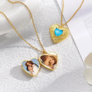 Two gold heart-shaped lockets displayed on a textured white surface, each hanging on a delicate gold chain. One locket is open, showing personalized couple photos inside, while another locket features an ornate gold design with a blue heart-shaped gem in the center. The arrangement highlights customizable romantic jewelry pendants.