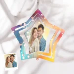 A hanging star-shaped personalized wind spinner ornament featuring a couple’s photo in the center. The star has layered cut-out edges with a multicolor gradient (blue, pink, orange, and yellow) and soft glowing light effects. It is suspended by a small chain and displayed against a light fabric background, with a small printed photo reference of the couple shown in the corner.