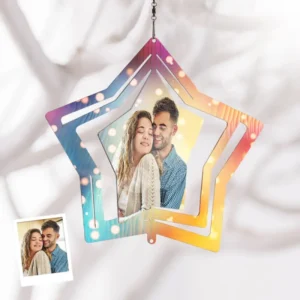 A hanging star-shaped personalized wind spinner ornament featuring a couple’s photo in the center. The star has layered cut-out edges with a multicolor gradient (blue, pink, orange, and yellow) and soft glowing light effects. It is suspended by a small chain and displayed against a light fabric background, with a small printed photo reference of the couple shown in the corner.