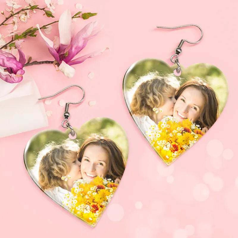 9bcd21cee8cd262158b438ce93009bb1webp - printshop4me.ae A pair of heart-shaped personalized photo earrings displayed on a pink background with floral decorations. Each earring contains a printed photo of a mother and child, featuring a metal hook and glossy heart frame design, suggesting custom photo jewelry gifts.