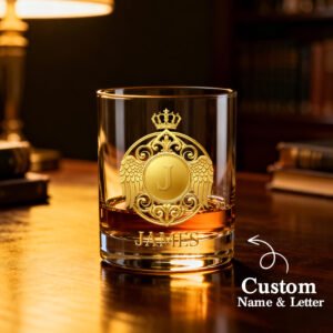 Personalized engraved whiskey glass featuring a custom monogram with an initial, name, crown, and winged crest design in a luxurious gold-style engraving. Displayed with whiskey on a wooden desk in a sophisticated setting, this custom rocks glass is a thoughtful gift for men, dads, husbands, groomsmen, birthdays, Father's Day, anniversaries, weddings, or Christmas.