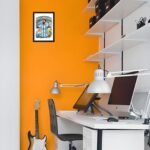 "Modern home office with orange accent wall featuring a framed Lionel Messi Argentina football artwork, desk setup with computer, lamp, and guitar in corner."
