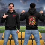 Morocco Supporter Hoodie - printshop4me.ae Morocco football men’s black hoodie worn in a stadium, featuring a small Morocco chest logo on the front and a large Morocco player graphic on the back with red and green flag-inspired design.