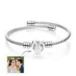 Silver twisted cable-style bracelet with a central heart-shaped charm featuring an engraved couple photo. The bracelet includes adjustable metal bead accents and is displayed on a clean white background, with a small reference image of the original couple photo shown in the bottom left corner.