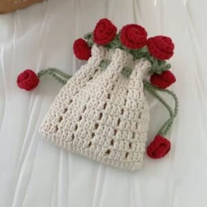 A handmade crochet bucket-style pouch in off-white yarn, decorated with green vine details and multiple red rose flowers attached around the top edge. The bag is displayed on a light fabric background and features a soft, textured knit design with floral embellishments, suggesting a handmade gift accessory or decorative pouch.