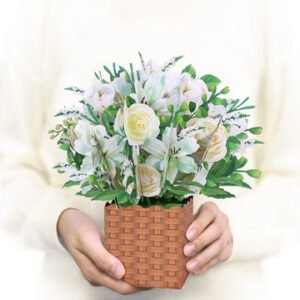 White floral arrangement in a woven basket featuring roses, artificial flowers, green leaves, and decorative foliage. A handmade flower bouquet gift display suitable for home decoration, celebrations, and special occasions.