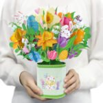 Easter floral pop-up greeting card featuring a colorful bouquet of spring flowers, decorated Easter eggs, green leaves, and cute bunny illustrations arranged in a paper flower pot. A festive 3D paper card for Easter greetings, seasonal gifting, and spring home decoration.