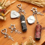 Set of personalized crystal keychains featuring engraved heart and oval lockets with the name "Emma," paired with decorative heart and star key rings, ribbon charms, and wire-wrapped crystal pendants in black and rose quartz. Displayed on a wooden tabletop with dried flowers, these custom name keychains make elegant personalized gifts.