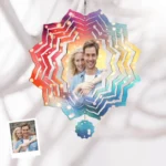 A hanging personalized wind-spinner style ornament featuring a couple’s photo in the center, surrounded by layered circular cut-out patterns in a rainbow gradient. The design creates a kaleidoscope-like effect with warm and cool colors radiating outward, suspended from a small chain, with a matching decorative charm below.