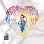 A hanging personalized heart-shaped wind spinner ornament featuring a couple’s photo in the center. The heart design is made of layered circular metal arcs with a multicolor gradient (pink, purple, blue, and gold tones) and soft glowing light effects. It is suspended from a small hook against a light fabric background, with a small reference photo of the couple shown in the corner.