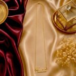 Gold cursive 'Hudda' name necklace on champagne satin with a gold jewelry box nearby.