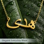 Gold necklace with Arabic-script pendant resting on a dew-covered green leaf.
