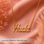Gold name necklace spelling 'Huda' on pink satin fabric with dried flowers, luxury jewelry.