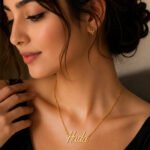 Close-up of a woman wearing a gold name necklace that reads 'Huda' with a matching gold flower stud earring worn on the ear.
