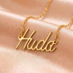 Gold name necklace spelling 'Huda' on pink satin fabric