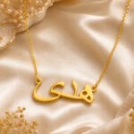 Gold name necklace with Arabic script pendant resting on pale satin fabric.